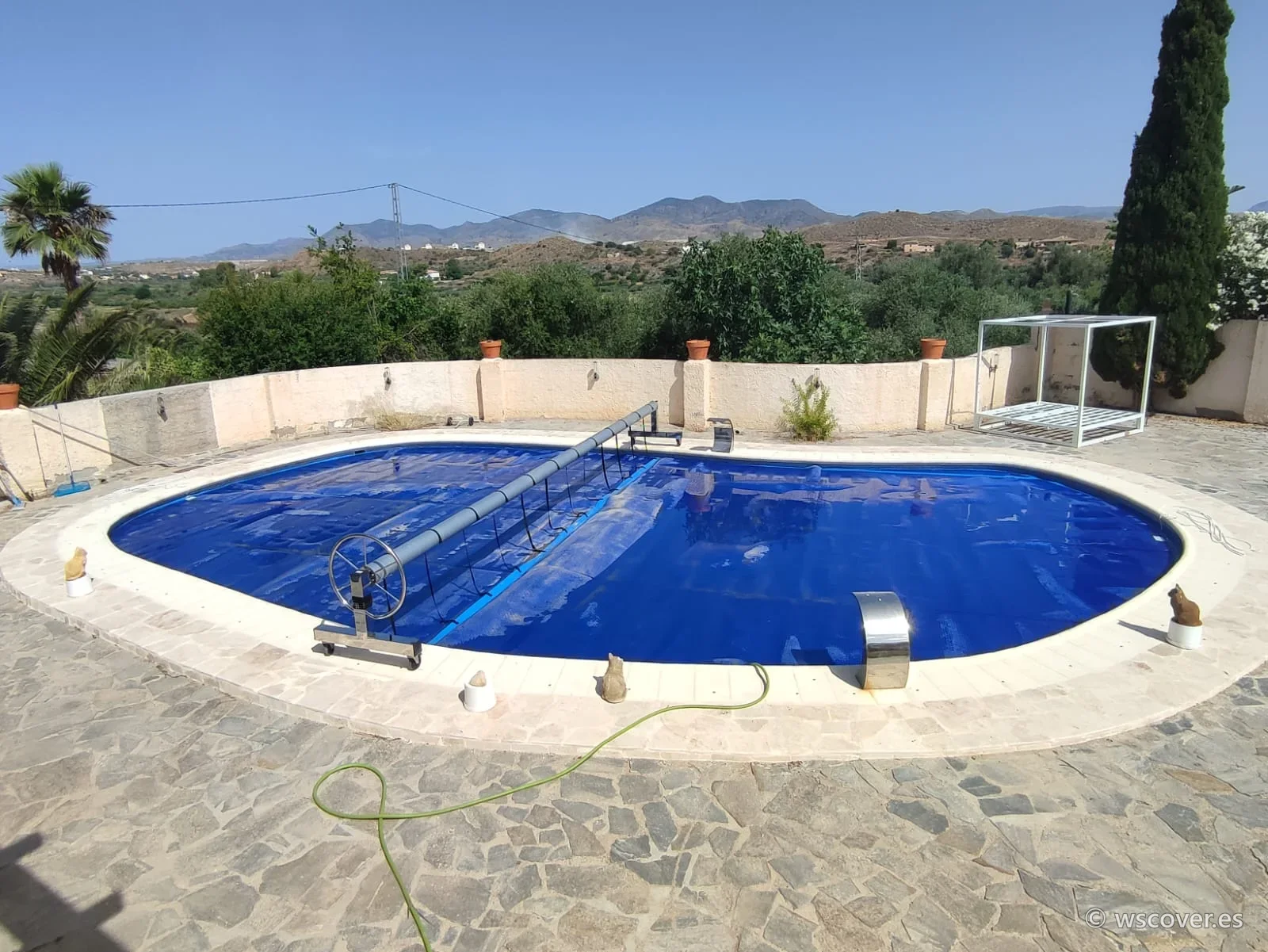 Cubierta de burbuja de 500 micras sobre piscina ovalada, con enrollador y la sierra al fondo